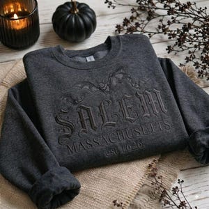 May include: A dark charcoal gray crewneck sweatshirt featuring black embroidered text that reads SALEM MASSACHUSETTS est 1626. Above the text, there is an embroidered design of two flying bats. The sweatshirt has long sleeves and a ribbed collar.