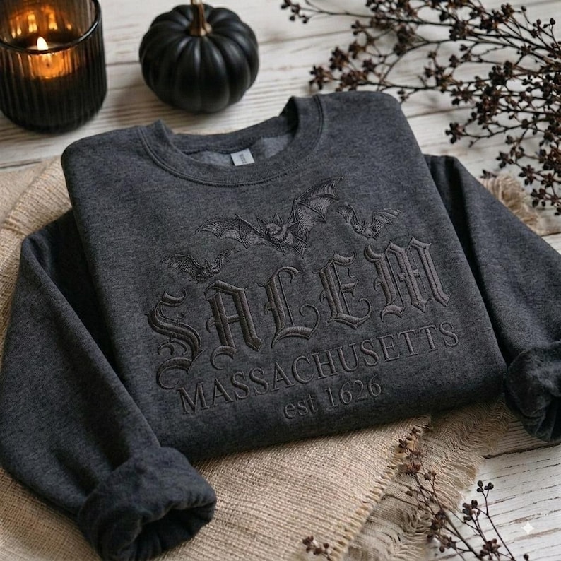 May include: A dark charcoal gray crewneck sweatshirt featuring black embroidered text that reads SALEM MASSACHUSETTS est 1626. Above the text, there is an embroidered design of two flying bats. The sweatshirt has long sleeves and a ribbed collar.