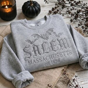 May include: A light gray crewneck sweatshirt featuring an embroidered design of bats and the text Salem Massachusetts est 1626 in a gothic font. The garment has long sleeves with ribbed cuffs and a ribbed neckline, showcasing a detailed stitched graphic.