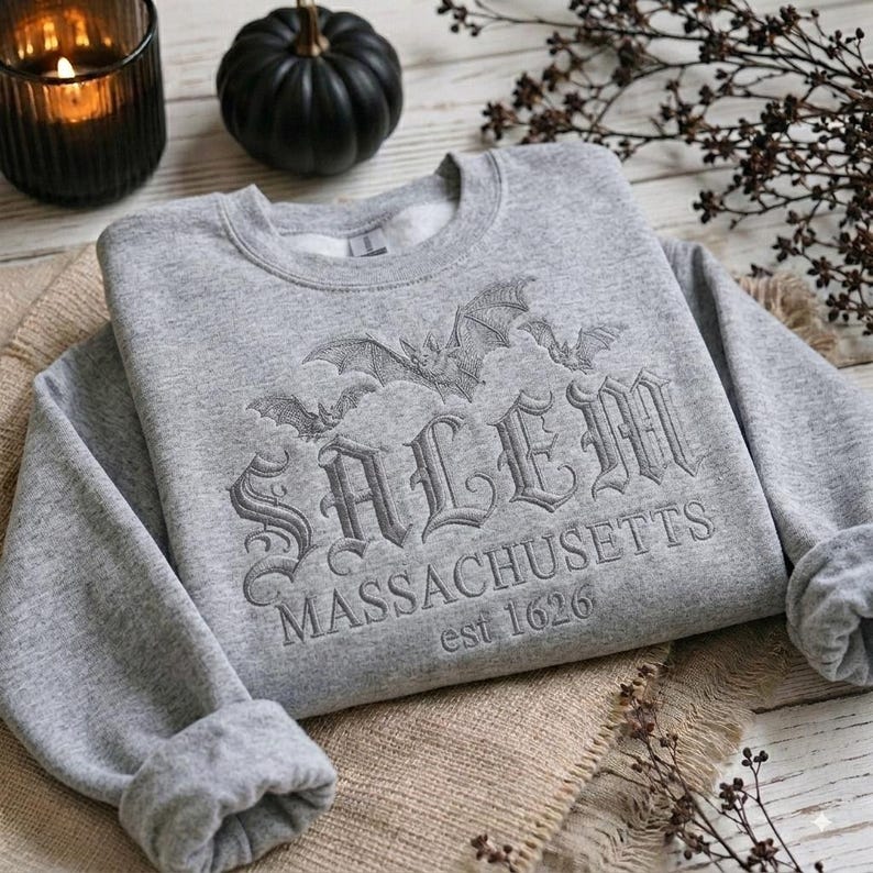 May include: A light gray crewneck sweatshirt featuring an embroidered design of bats and the text Salem Massachusetts est 1626 in a gothic font. The garment has long sleeves with ribbed cuffs and a ribbed neckline, showcasing a detailed stitched graphic.