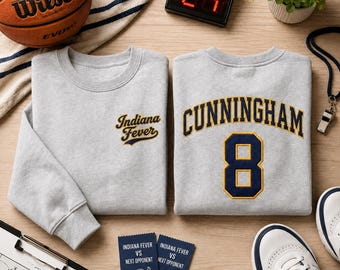 Puede incluir: Dos sudaderas de cuello redondo en color gris claro. La sudadera de la izquierda presenta el texto Indiana Fever bordado en azul marino y dorado. La de la derecha muestra el nombre CUNNINGHAM y el número 8 en letras azul marino y doradas.