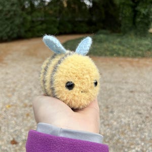May include: A crocheted bee plush toy with yellow and black stripes, blue wings, and black eyes. The bee is being held in a hand wearing a purple jacket.