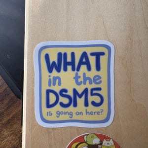 Puede incluir: Una pegatina cuadrada de esquinas redondeadas con fondo amarillo p&aacute;lido y borde azul. El texto "WHAT in the DSM5 is going on here?" aparece en letras azules sin remates. Debajo hay una pegatina circular con varios personajes tipo sushi.