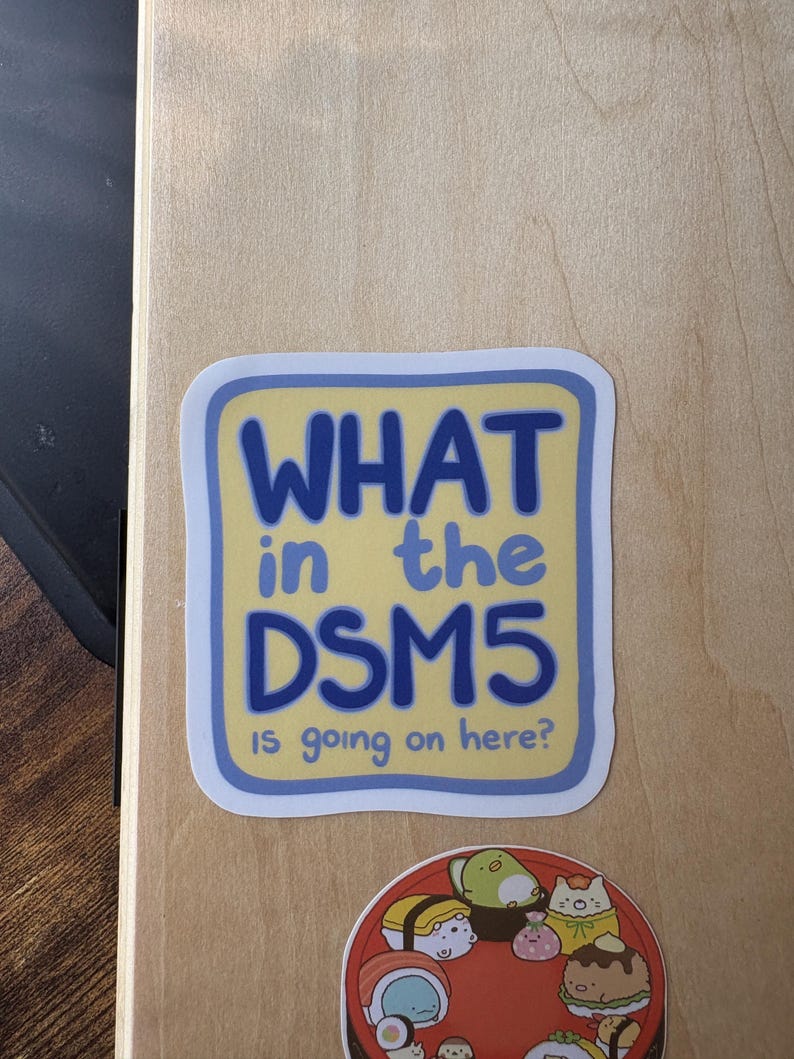 Puede incluir: Una pegatina cuadrada de esquinas redondeadas con fondo amarillo p&aacute;lido y borde azul. El texto "WHAT in the DSM5 is going on here?" aparece en letras azules sin remates. Debajo hay una pegatina circular con varios personajes tipo sushi.