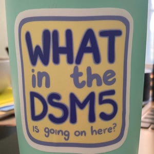 Puede incluir: Un vaso de acero inoxidable de color verde azulado claro con borde y base plateados. Una pegatina rectangular en el frente presenta un fondo amarillo con un borde morado y el texto "WHAT in the DSM5 IS going on here?" en color azul.