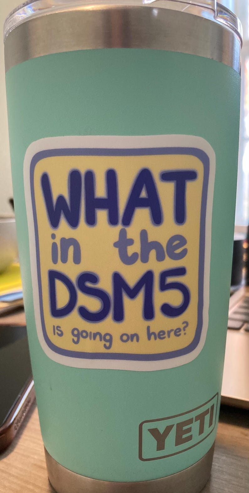 Puede incluir: Un vaso de acero inoxidable de color verde azulado claro con borde y base plateados. Una pegatina rectangular en el frente presenta un fondo amarillo con un borde morado y el texto "WHAT in the DSM5 IS going on here?" en color azul.