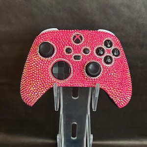 May include: A pink Xbox controller covered in sparkling rhinestones. The controller is on a clear plastic stand.
