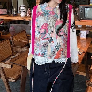 May include: A vibrant bird-patterned vest in pink, blue, and green, with a bright pink trim. It's worn over a white top with lace detail. The model is wearing dark blue jeans. The overall style is a mix of patterns and textures.