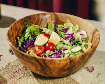 May include: A round wooden bowl with a distinct grain pattern filled with a fresh salad containing chopped green lettuce, shredded purple cabbage, sliced cucumbers, red cherry tomatoes, a tomato slice, and two yellow lime wedges.