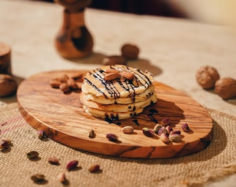 May include: A stack of pancakes topped with chocolate sauce and almonds, served on a wooden cutting board with pistachios scattered around.