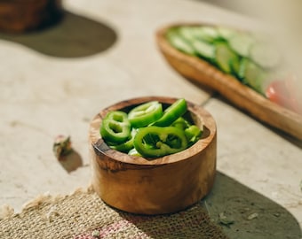 May include: A small wooden bowl filled with sliced green peppers, sitting on a burlap placemat. The bowl is made of olive wood and has a natural, rustic finish.