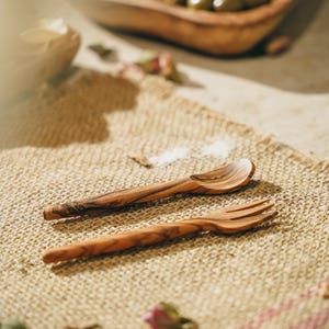 Könnte beinhalten: Zwei Holzutensilien, ein Löffel und eine Gabel, liegen auf einer Jutestoff-Oberfläche. Die Utensilien sind aus Olivenholz gefertigt und haben eine natürliche, rustikale Oberfläche.