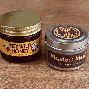 May include: Two containers on a wooden surface. One is a glass jar of Feywild Honey with a gold lid and a label featuring a bee and a die. The other is a silver tin of Meadow Mead Natural Soy Wax Candle with a tree of life design.