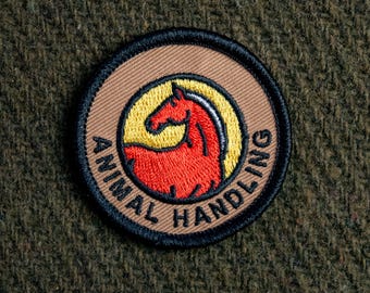 Puede incluir: Un parche bordado marrón y negro con una silueta de caballo rojo dentro de un círculo amarillo. El texto "ANIMAL HANDLING" está escrito alrededor del círculo.