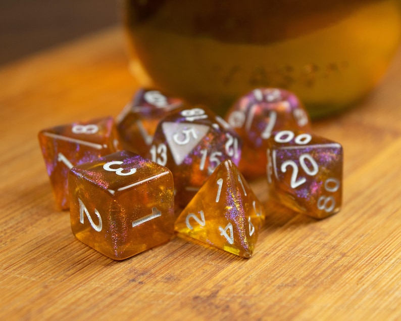 May include: A collection of seven amber-colored polyhedral dice with white numbering. The dice have a shimmering, glittery effect with purple and pink accents. The dice are arranged on a wooden surface.