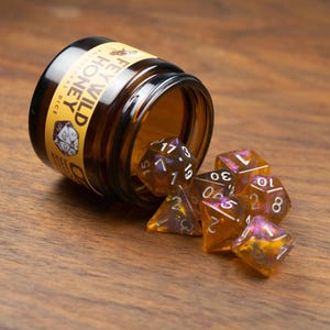May include: A collection of polyhedral dice spilling from a small brown glass jar. The dice are transparent with orange and purple hues, and white numbering. The jar has a yellow label with the text "FEYWILD HONEY".