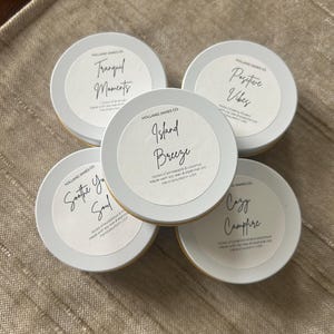 Puede incluir: Cinco pequeños botes de velas redondos plateados con etiquetas blancas. Las etiquetas presentan texto escrito a mano: "Tranquil Moments", "Positive Vibes", "Island Breeze", "Soothe Your Soul" y "Cozy Campfire". Las velas están sobre una superficie texturizada de tono neutro.