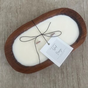 May include: A cream-colored candle in a dark brown wooden bowl. The candle has two wooden wicks and is tied with black string. A white tag with the words "Island Breeze" is attached.