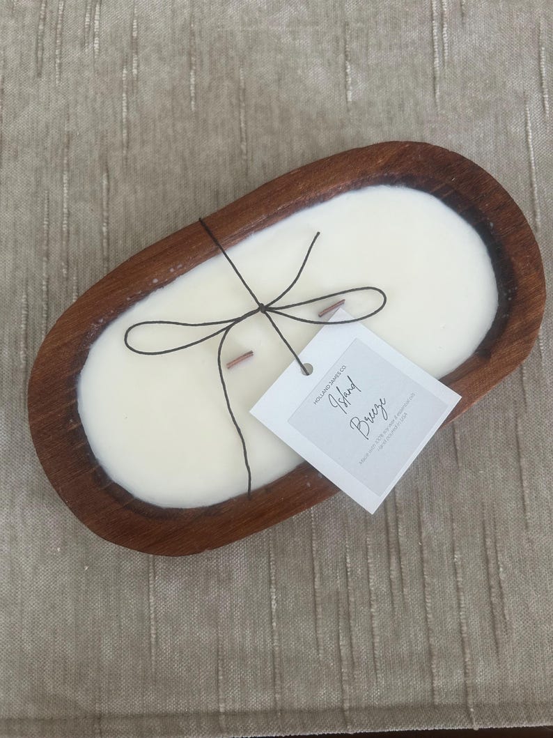 May include: A cream-colored candle in a dark brown wooden bowl. The candle has two wooden wicks and is tied with black string. A white tag with the words "Island Breeze" is attached.