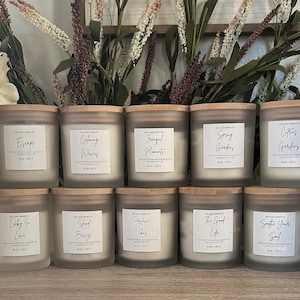 May include: A collection of 12 frosted glass candle jars with wooden lids. Each jar has a different label with a handwritten font and a unique scent name, such as "Date Night", "Escape", "Morning Madness", and "Cozy Campfire".
