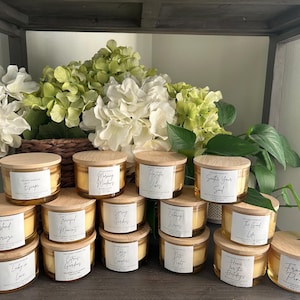 May include: A collection of 16 amber glass candle jars with wooden lids. The candles are arranged in a pyramid shape on a wooden shelf. The candles have various scents, including "Escape", "Morning Mimosas", "Positive Vibes", "Soothe Your Soul", "Island Breeze", "Fragrant Memories", "Spring Garden", "Ocean Waves", "Cozy Campfire", "Date Night", "Citrus Garden", "Home for the Holidays", and "Frosted Pines".