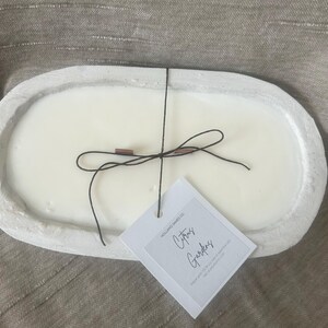 May include: A white oval-shaped candle in a concrete-like container. The candle has a black string tied around it with a small tag that reads "Citrus Gardens". The candle is on a neutral-colored surface.