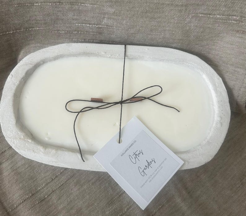 May include: A white oval-shaped candle in a concrete-like container. The candle has a black string tied around it with a small tag that reads "Citrus Gardens". The candle is on a neutral-colored surface.