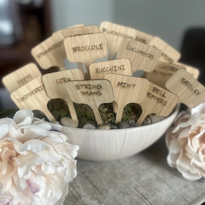 May include: A collection of wooden garden markers, each labeled with the names of various vegetables and herbs, including "Broccoli," "Zucchini," and "Mint." The markers are displayed in a white bowl filled with moss and small stones, with a blurred background.
