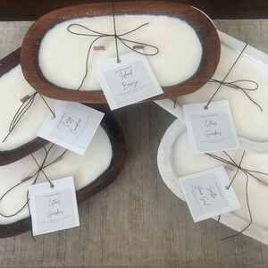 May include: Several oval-shaped candles in wooden and white containers. Each candle has a white wax surface, a wooden wick, and a tag with a scent name. The candles are arranged on a neutral-colored surface.