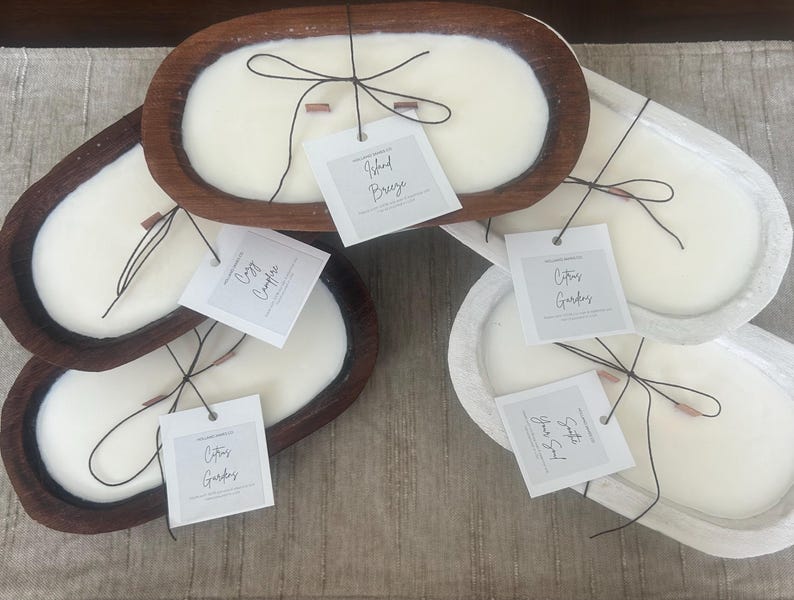 May include: Several oval-shaped candles in wooden and white containers. Each candle has a white wax surface, a wooden wick, and a tag with a scent name. The candles are arranged on a neutral-colored surface.