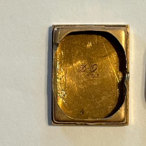 K&ouml;nnte beinhalten: Ein rechteckiges, goldfarbenes Uhrengeh&auml;use mit abgenutztem, strukturiertem Innenraum. Im Inneren des Geh&auml;uses befinden sich ge&auml;tzte Markierungen, darunter die Buchstaben "D-O" und "R.G.P.". Das Geh&auml;use hat eine leicht gebogene Form und einen sichtbaren Rahmen.