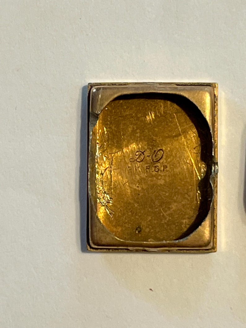 K&ouml;nnte beinhalten: Ein rechteckiges, goldfarbenes Uhrengeh&auml;use mit abgenutztem, strukturiertem Innenraum. Im Inneren des Geh&auml;uses befinden sich ge&auml;tzte Markierungen, darunter die Buchstaben "D-O" und "R.G.P.". Das Geh&auml;use hat eine leicht gebogene Form und einen sichtbaren Rahmen.
