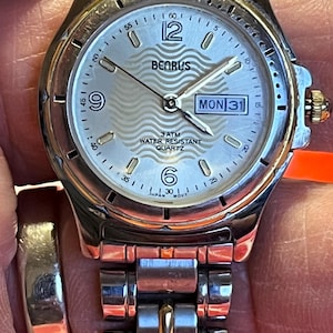 Op de afbeelding: Een zilverkleurig Benrus quartz polshorloge met een lichtgekleurde wijzerplaat. Het horloge heeft een datumweergave, een secondewijzer en een zilverkleurige band. De wijzerplaat toont de tekst "BENRUS", "3 ATM WATER RESISTANT QUARTZ" en "JAPAN MOVT".