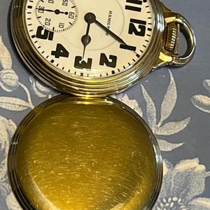May include: A gold-coloured pocket watch with a white face and black numerals. The watch face displays the brand name "HAMILTON". The watch has a second dial and a gold-coloured case. The watch is open, revealing the back of the case.
