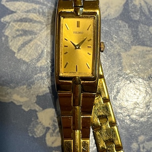 May include: A gold-toned Seiko wristwatch with a rectangular face and a matching link band. The watch face displays the Seiko logo and the words "MOV'T JAPAN". The watch has a classic, elegant design.