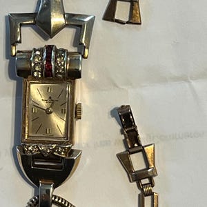 Może przedstawiać: Zegarek na rękę w stylu vintage w kolorze złotym z prostokątną tarczą i uszkodzonym paskiem. Tarcza zegarka ma złote wskazówki i znaczniki, z czerwonym i przezroczystym kamieniem. Pasek jest częściowo odłączony.