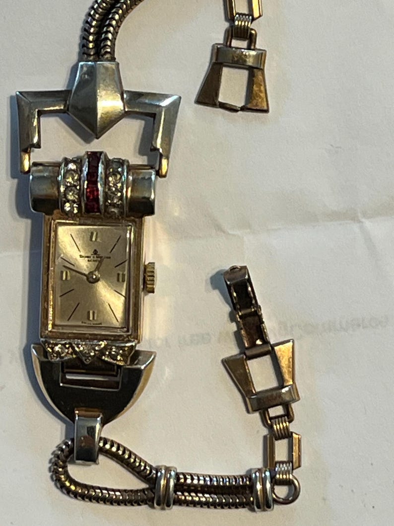 K&ouml;nnte beinhalten: Eine goldfarbene Vintage-Armbanduhr mit rechteckigem Zifferblatt und defektem Armband. Das Zifferblatt hat goldene Zeiger und Markierungen, mit einem roten und klaren Steinakzent. Das Armband ist teilweise abgetrennt.