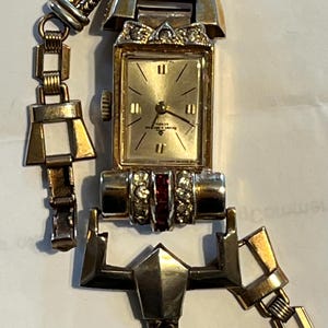 K&ouml;nnte beinhalten: Vintage goldfarbene Armbanduhr mit rechteckigem Zifferblatt und goldenem Armband. Das Zifferblatt ist goldfarben mit schwarzen Ziffern und Zeigern. Das Armband hat geometrische und Kettenglieder-Designs mit roten und klaren Steinen.