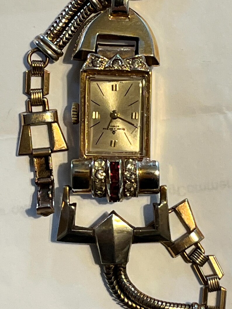 K&ouml;nnte beinhalten: Vintage goldfarbene Armbanduhr mit rechteckigem Zifferblatt und goldenem Armband. Das Zifferblatt ist goldfarben mit schwarzen Ziffern und Zeigern. Das Armband hat geometrische und Kettenglieder-Designs mit roten und klaren Steinen.