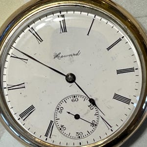 Könnte beinhalten: Eine goldene Taschenuhr mit römischen Ziffern für die Stunden und einem kleineren Hilfszifferblatt für die Sekunden. Das Zifferblatt der Uhr ist weiß mit schwarzen Markierungen und dem Wort "Howard" in Schwarz gedruckt.