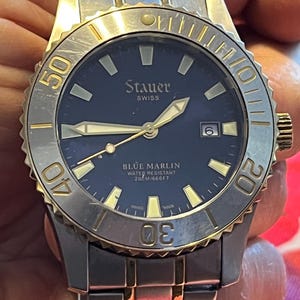 May include: A stainless steel and gold-toned Stauer Blue Marlin watch with a dark blue face. The watch features gold-toned hands and markers, a date display, and a rotating bezel. The watch is water resistant to 660 feet.