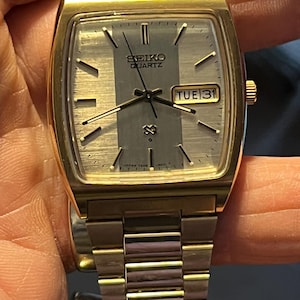 May include: A gold-toned Seiko Quartz watch with a rectangular face. The watch face is silver with a vertical gray stripe and displays the day and date. The watch band is gold-toned, and the watch is held by a hand.