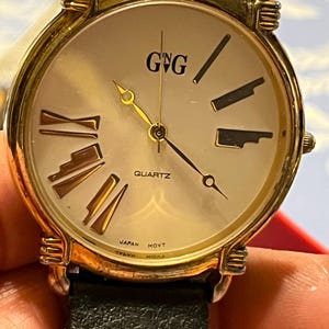 May include: A gold-toned quartz watch with a cream-coloured face, featuring gold Roman numerals and black markers. The watch has a black leather strap and the text "GNG" and "QUARTZ" are visible on the face. The watch is marked "JAPAN MOVT".