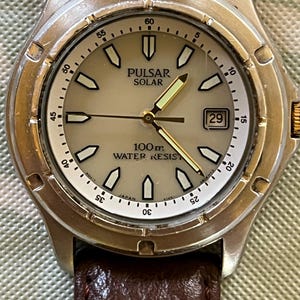 May include: Silver Pulsar Solar watch with a light-coloured face, black and white markers, and gold hands. The watch has a brown leather strap and a date display at the 3 o'clock position. Water-resistant to 100m.