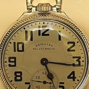 May include: A close-up of a vintage Hamilton Railway Special pocket watch. The watch has a cream face with black numerals and hands. A smaller seconds dial is at the bottom. The watch is encased in gold-toned metal with a loop at the top.