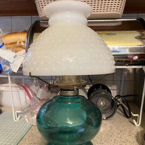 Op de afbeelding: Een vintage olielamp met een witte, gematteerde glazen kap en een tealblauwe glazen voet. De kap heeft een getextureerd, gebolde oppervlak.