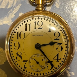 May include: A gold-coloured Waltham pocket watch with a cream face and black numerals. The watch features a subdial and a textured crown. The hands are black, and it has a gold-coloured loop for suspension.