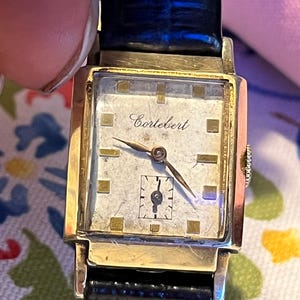 May include: A vintage gold-toned Cortébert wristwatch with a rectangular face and a black leather band. The watch face is off-white with gold-toned markers and hands. The brand name "Cortébert" is printed on the watch face.