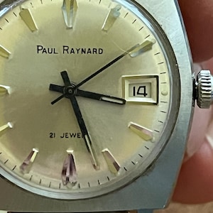 May include: A vintage Paul Raynard wristwatch with a silver-colored case and a gold-toned face. The watch features black hands, a date window displaying the number 14, and the text "21 Jewels". The watch has a textured crown and a gold-colored band.