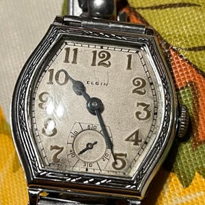 Op de afbeelding: Vintage Elgin polshorloge met een zilverkleurige achthoekige kast. De wijzerplaat is gebroken wit met zwarte cijfers en wijzers. Een kleinere subdial bevindt zich onderaan. De horlogeband is zilverkleurig.